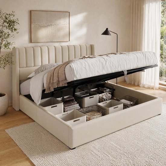 Lit beige rembourré avec rangement hydraulique ouvert, rempli de paniers, de couvertures et de coussins. Visible dans une chambre lumineuse avec une fenêtre.