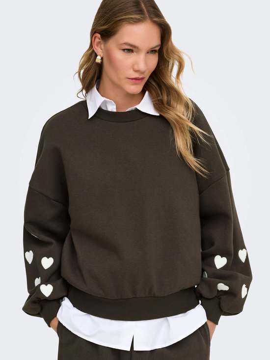 Donkerbruine sweater met witte hartjes op de mouwen, gedragen door een model met een witte blouse eronder.