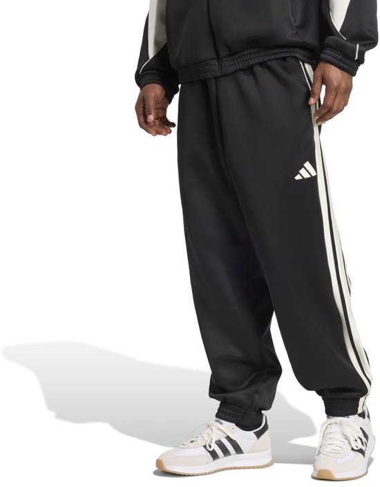 Man in zwarte trainingsbroek met witte strepen en Adidas logo, gedragen met witte sneakers.