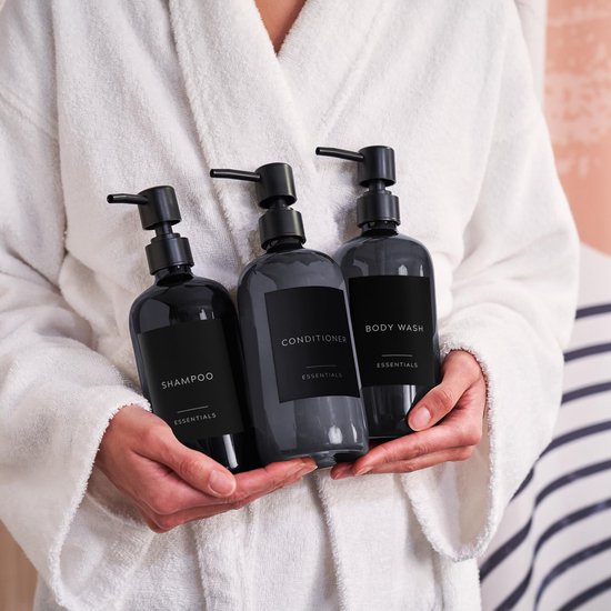 Iemand in een witte badjas houdt drie zwarte pompdispensers vast. Labels: SHAMPOO, CONDITIONER, BODY WASH.