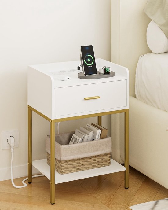 Wit nachtkastje met gouden pootjes, lade en plank, naast een bed, met telefoon die draadloos oplaadt en een mand met boeken.