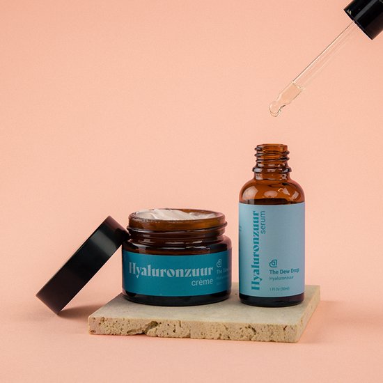 Glazen flesje Hyaluronzuur serum en potje crème, op een stenen plateau met roze achtergrond. Een druppelaar met serum erboven.