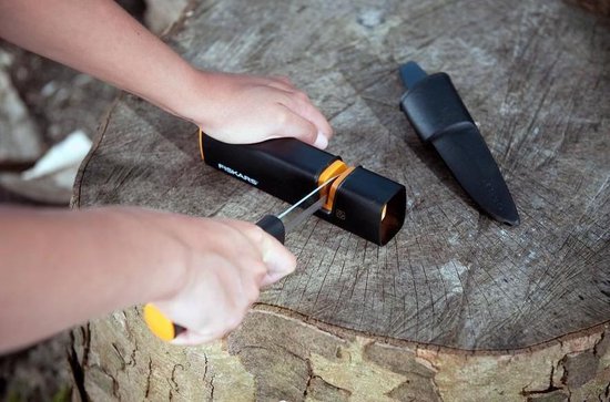 Handen gebruiken een zwarte Fiskars messenslijper met oranje accenten op een houten stronk.