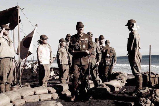 Groep militairen op een strand bij zandzakken. Man in midden draagt een donker uniform met medailles.