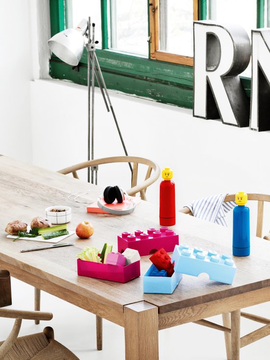 Houten tafel met roze en blauwe LEGO lunchboxen, gevuld met eten. Rode en blauwe LEGO flessen met LEGO hoofden.