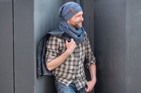 Man met blauwe beanie en sjaal, draagt geruite blouse en donkere jas over schouder.
