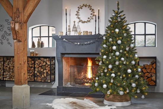 Sfeervolle kerstboom met zilveren ornamenten naast een brandende open haard in een woonkamer.
