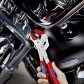 Hand houdt een tang met rode en blauwe handvatten vast, die een deel van een motorfiets aanpast.