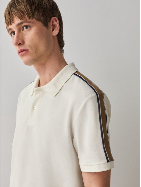 Close-up van een man met kort bruin haar die een crème poloshirt met strepen op de schouders draagt.