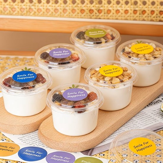 Zes kleine potjes met yoghurt, granola en gedroogd fruit, elk met een etiket 'Smile For Happiness'.