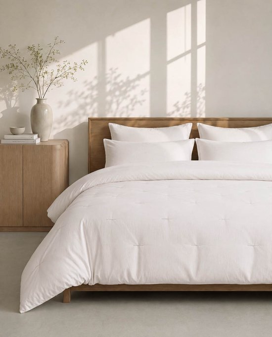 Wit gestikt dekbed op een houten bed met houten nachtkastje en vaas met takken.