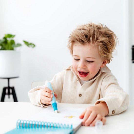 Jongen met blond haar die in een notitieboekje tekent met een blauw-witte pen, hij draagt een crème kleurig shirt.