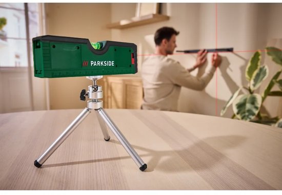 Groene Parkside laserwaterpas projecteert rode lijnen op een muur terwijl een man een schilderij ophangt.