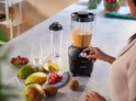 Zwarte Philips blender met een oranje smoothie in de kan, bediend door een vrouw, met fruit zoals aardbeien, mango en kiwi.