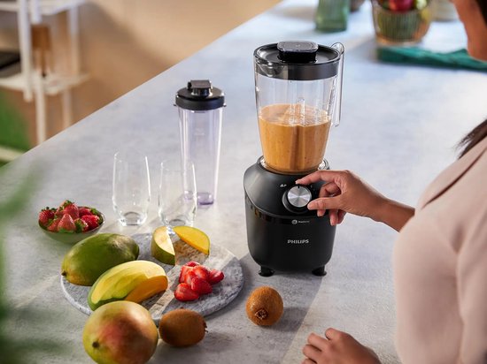 Zwarte Philips blender met een oranje smoothie in de kan, bediend door een vrouw, met fruit zoals aardbeien, mango en kiwi.