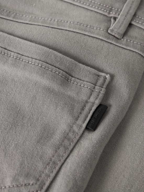 Detail van een grijze jeans met een opgestikte zak en een zwart logolabel aan de zijkant.