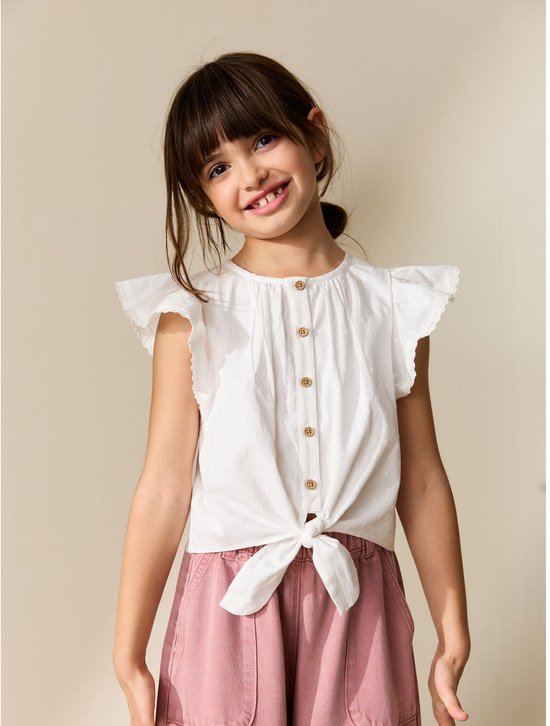 Meisje draagt een witte blouse met ruches, houten knopen en gestrikte voorkant, gecombineerd met een roze broek.