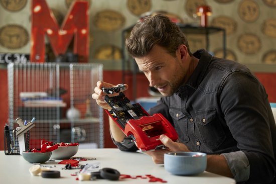 Een man zit aan tafel en monteert een rode LEGO Chevrolet Corvette cabriolet. Onderdelen liggen verspreid op tafel.