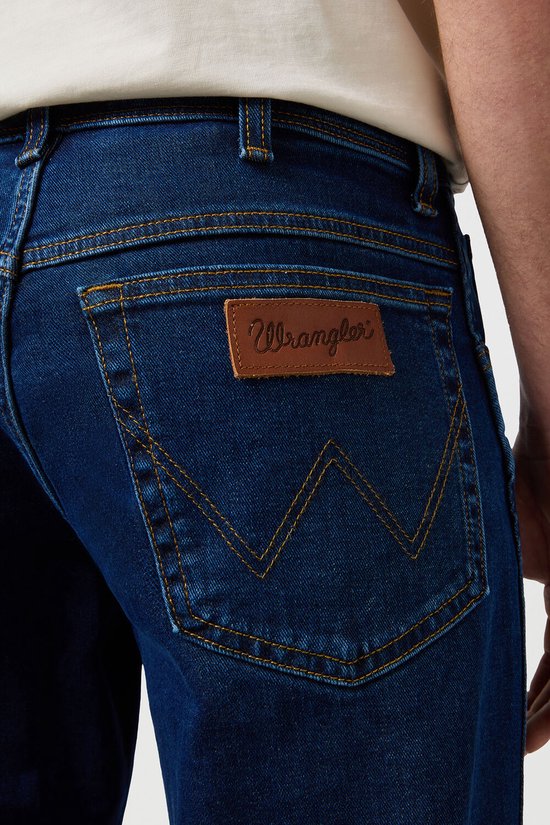 Donkerblauwe Wrangler jeans met bruin lederen logo op de achterzak en kenmerkend stiksel. Gecropte foto van de achterkant.
