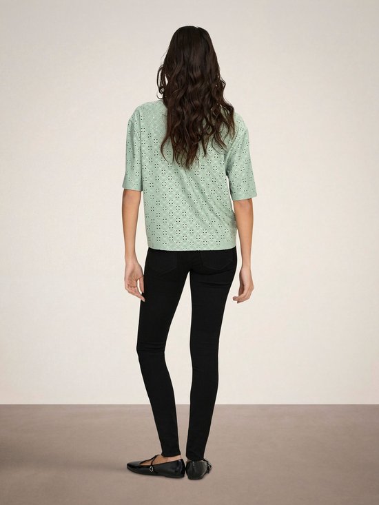Model met gekruiste armen in een lichtgroene blouse met broderie anglaise, korte mouwen en knopen.
