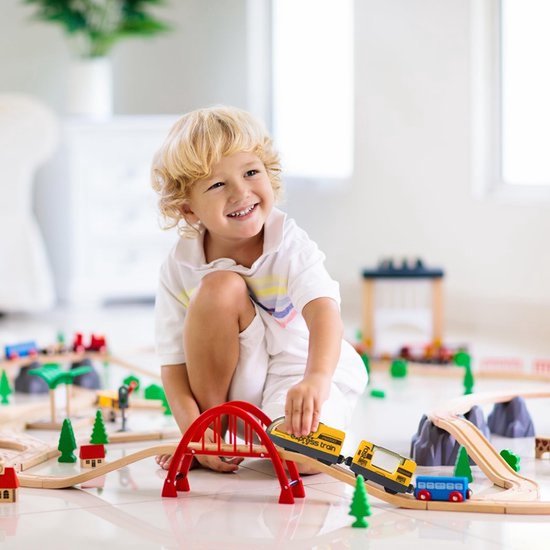 Een lachende jongen speelt met een gele trein op houten rails met een rode brug en berg.