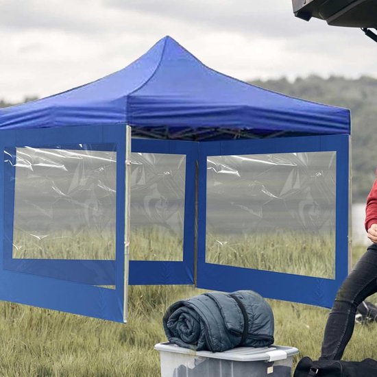 Blauwe partytent met transparante vensters, opgesteld in een open veld met een man die een kampeerbrander vasthoudt.