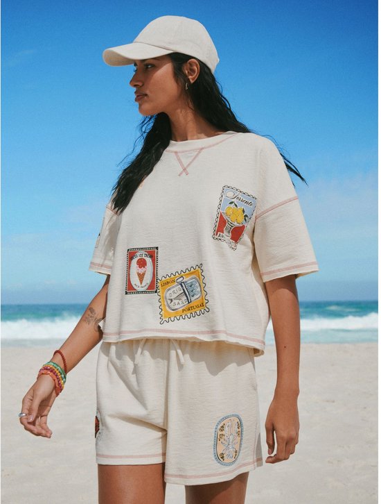 Vrouw in crème T-shirt en short met gestikte vakantie-emblemen en een pet op het strand.