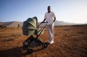 Groene kinderwagen met reiswieg, naast een man in een lichtgekleurde outfit, in een woestijnlandschap.