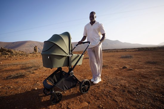 Groene kinderwagen met reiswieg, naast een man in een lichtgekleurde outfit, in een woestijnlandschap.