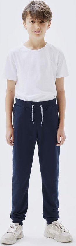 Jongen met donkerblauwe joggingbroek, witte t-shirt met korte mouwen en witte sneakers.
