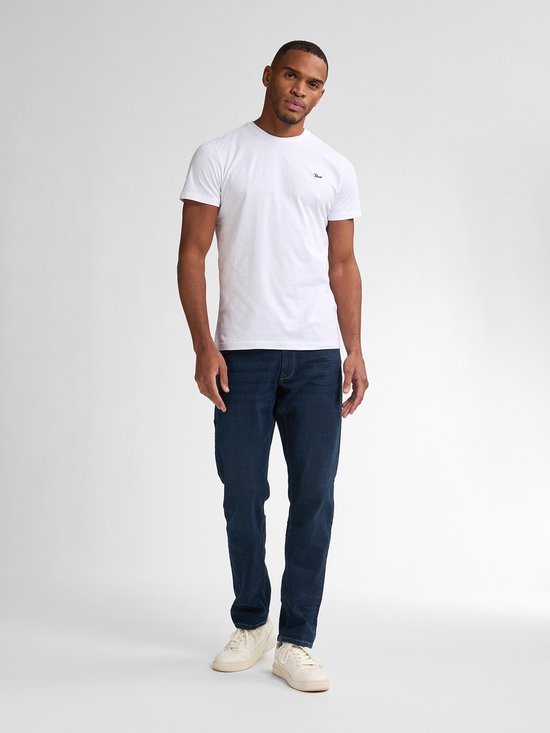 Model in een wit T-shirt en donkerblauwe tapered fit jeans tegen een lichte achtergrond.