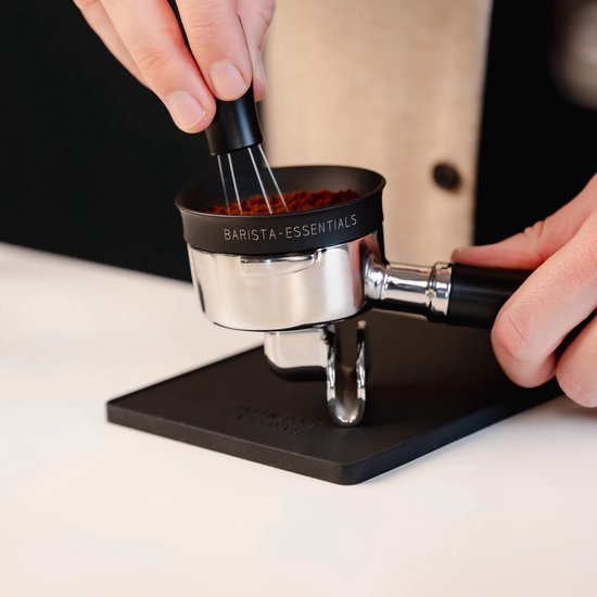 Een portafilter met gemalen koffie en een tamper-tool, staand op een zwarte tampermat.