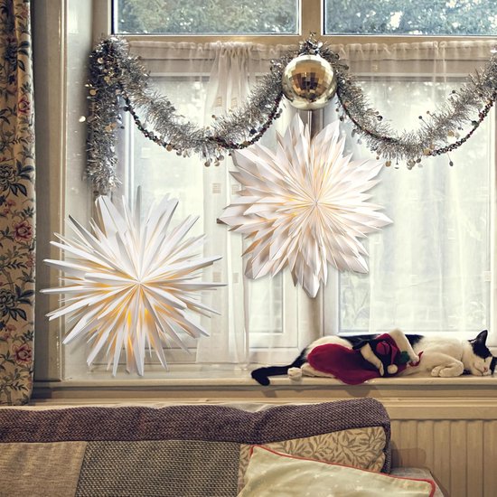 Twee witte papieren sterren met warm licht hangend voor een raam met kerstdecoratie en slapende kat.