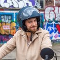 Man met donkerblauwe helm met vizier en beige jas, zittend op een scooter.