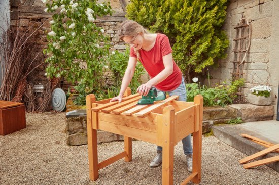 Vrouw schuurt verhoogde houten plantenbak met groene Bosch multischuurmachine, stofopvangbak bevestigd.
