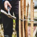 Hand met hamer slaat paal van houten schutting aan. Palen verbonden met metaaldraad.