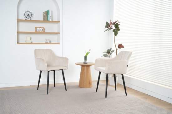 Twee crème fluwelen eetkamerstoelen met zwarte poten, rond een houten tafel met bloemen.