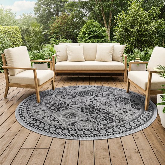 Rond grijs en zwart vloerkleed met mozaïekpatroon op een houten terras met buitenmeubilair.