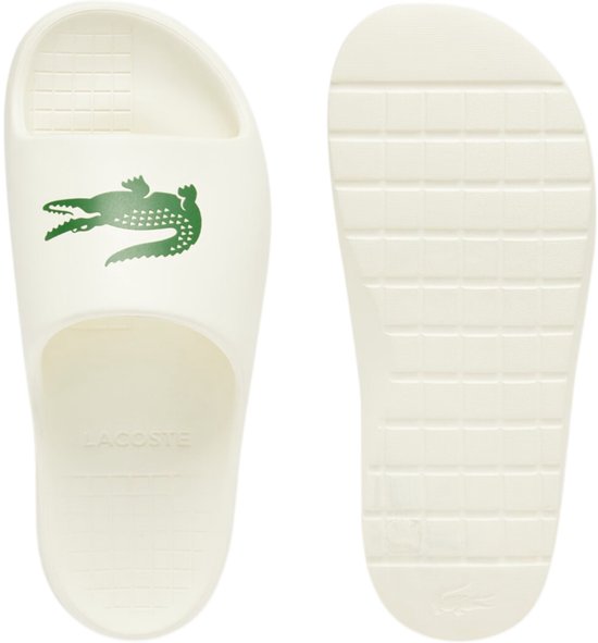 Crèmekleurige Lacoste slipper, met groen krokodillenlogo en 'LACOSTE' op de zool. Onderkant van de slipper zichtbaar.