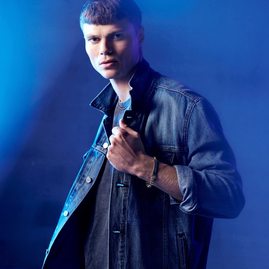 Man in denim jack draagt zilverkleurige ID-plaatarmband, verlicht door blauw licht.