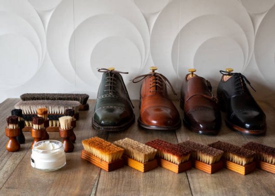 Vijf paren schoenen, borstels en een potje witte schoencrème op een houten ondergrond.