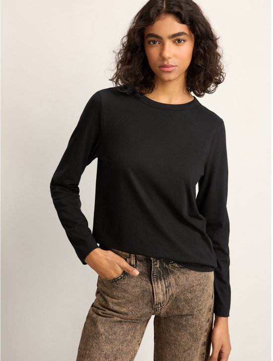 Vrouw met krullend haar, hand in broekzak, draagt een zwarte longsleeve.