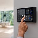 Een hand raakt het scherm aan van een zwart Shelly Wall Display XL, gemonteerd op een muur in een moderne woonkamer.