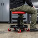 Rolkruk met rode zitting en twee zwarte opbergplateaus. Een man met groene werkbroek zit erop, met gereedschapskasten op de achtergrond.