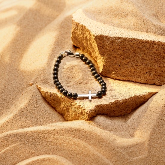 Zwarte onyx kralenarmband met zilverkleurig kruisdetail, liggend op zand.