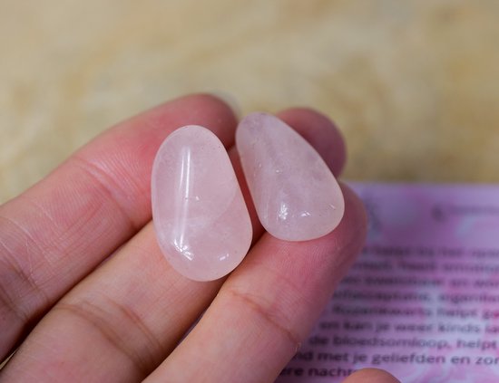 Een hand houdt twee gladde, lichtroze rozenkwarts edelstenen vast. De ene is ovaal, de andere druppelvormig.