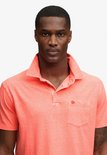 Close-up van een mannelijk model in een koraalrood, gemêleerd poloshirt met borstzak.