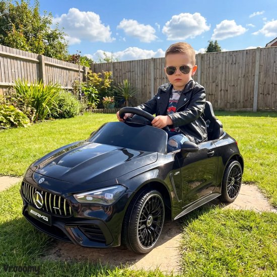 Jongetje in zwart leren jack en zonnebril rijdt in een zwarte Mercedes-Benz kinderauto in de tuin.