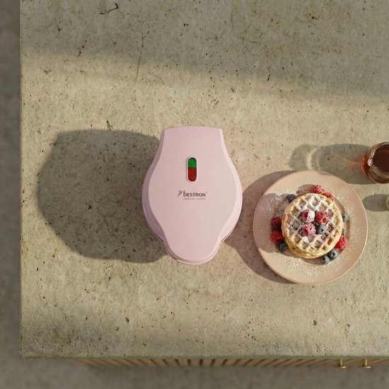 Roze Bestron wafelijzer gesloten, naast een stapel wafels met fruit en een potje honing.