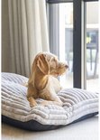 Een lichtbruine hond ligt op een grijs, ribbed hondenkussen bij een raam.